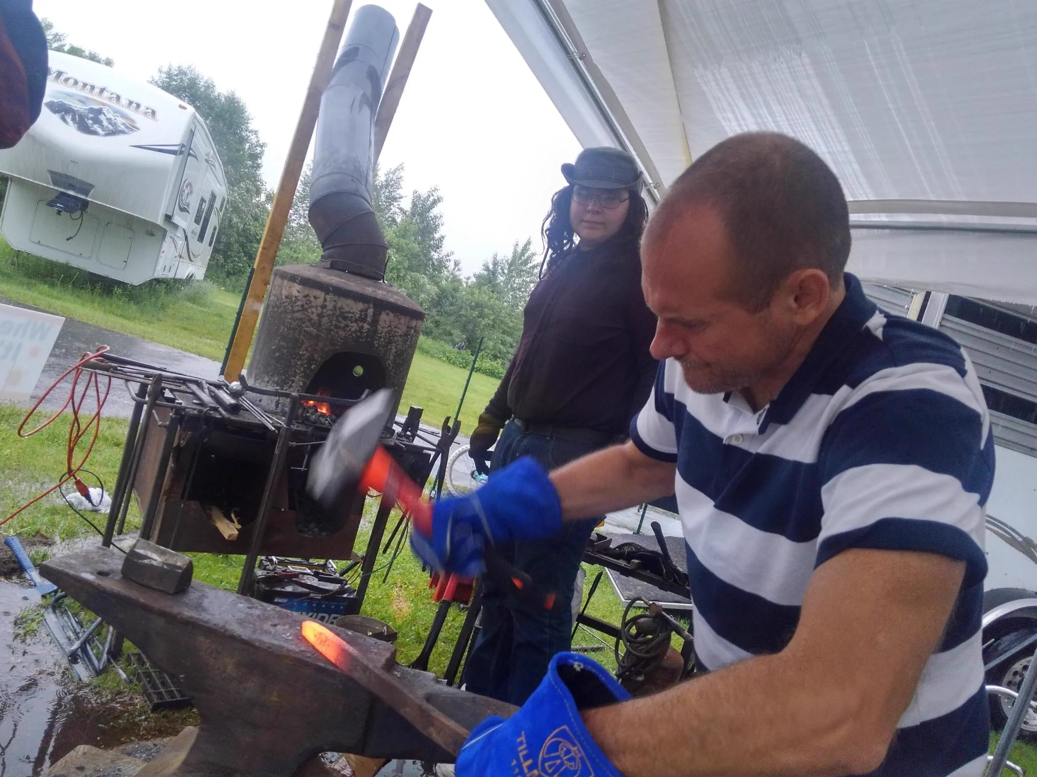Ernie, a volunteer blacksmith mentor, holding glowing hot iron with tongs on an anvil, with a coal forge behind him