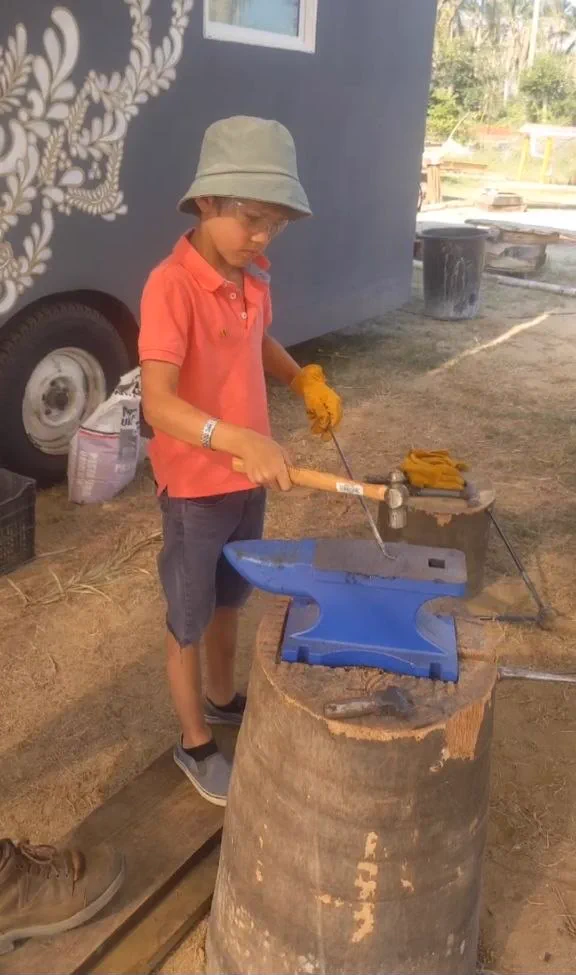 A young blacksmithing student in safety glasses and leather gloves, hammer in hand, working hot iron on a blue anvil