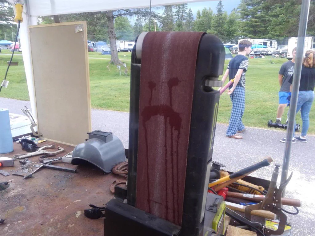 A belt sander station at Mancamp 2018 with kids working at the grinder under a tent
