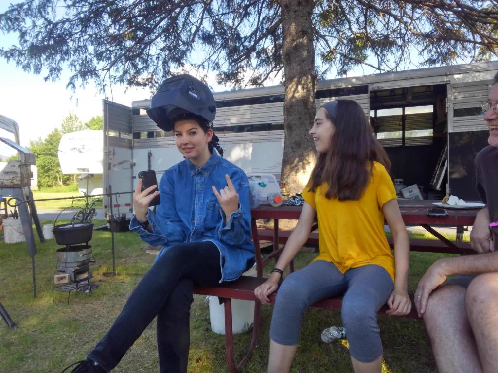 Two campers at Mancamp 2018, one wearing a welding helmet, hanging out by the trailer at Porcfest
