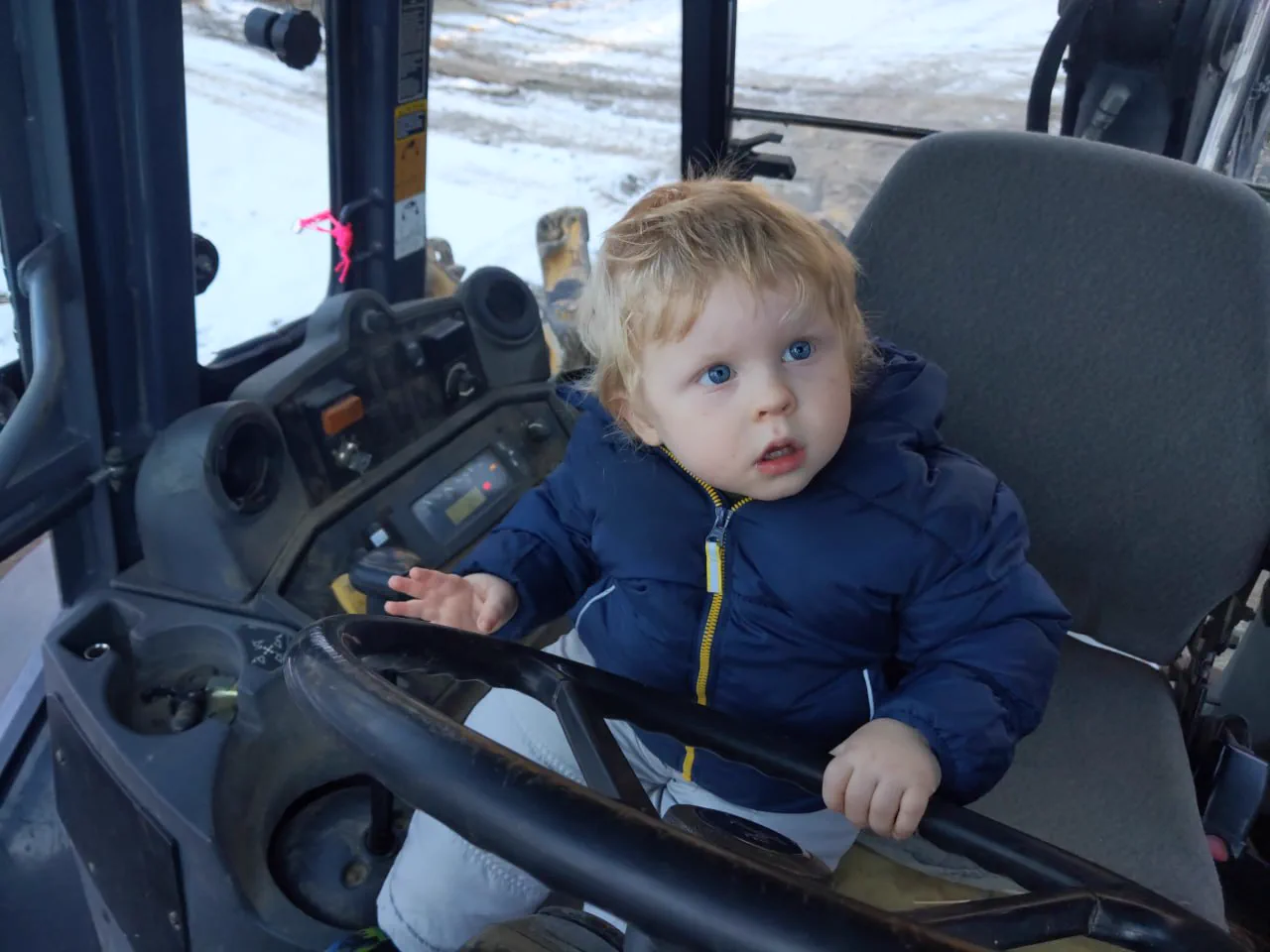 A young child with curly blond hair sitting at the controls of a tractor cab, hands on the steering wheel